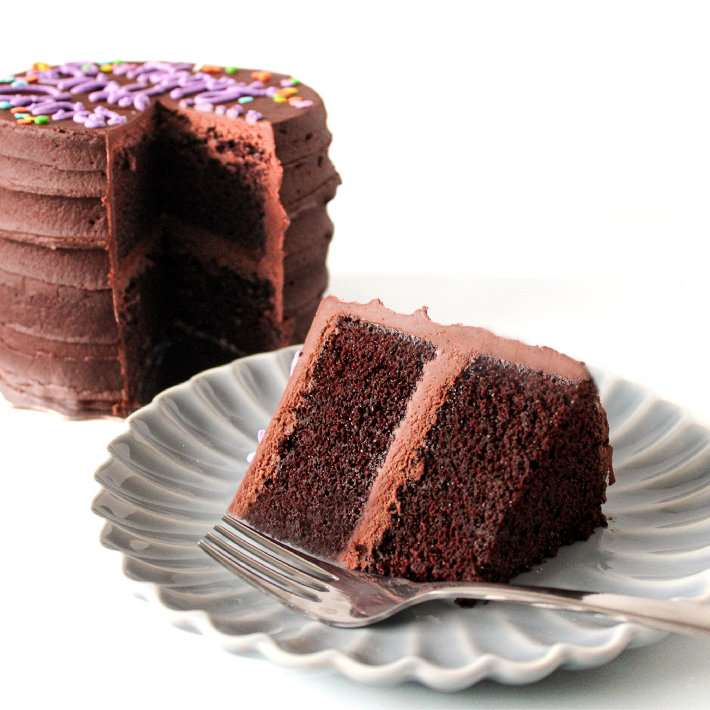 A slice of chocolate cake on a plate with a fork, next to a small chocolate cake with chocolate buttercream icing
